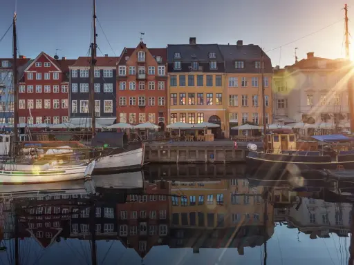 Färgglada hus vid en kanal i Köpenhamn, med båtar i förgrunden. Solen skiner och reflekteras i vattnet. En perfekt vy för en flyg till Köpenhamn-resa.