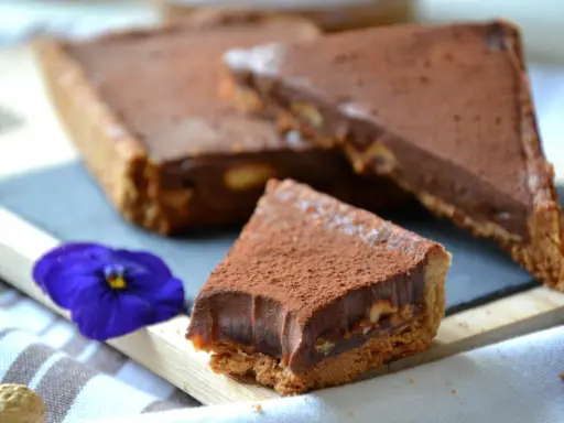 Tranche de tarte façon snickers, garnie de chocolat et de cacahuètes, saupoudrée de cacao. Une fleur bleue décore la présentation.
