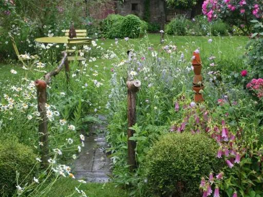 Un charmant jardin de curé, sauvage et fleuri, avec un chemin de dalles mouillées, des marguerites, des campanules et une table de bistrot.