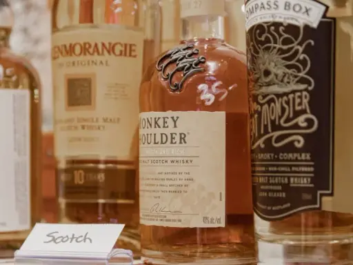 Flera flaskor whisky, inklusive Monkey Shoulder och Glenmorangie, står uppradade. En liten skylt med texten "Scotch" syns framför.