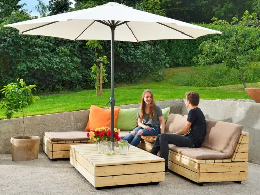 Två personer njuter av solen under ett parasoll vid en hemmabyggd loungesoffa i trä.