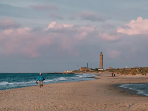 En person promenerar på stranden vid Skagens fyr. Molnen färgas rosa i solnedgången.