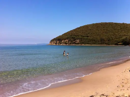 Spiaggia della Maremma con due bambini che giocano nell'acqua cristallina, sotto un cielo azzurro e una collina verdeggiante.
