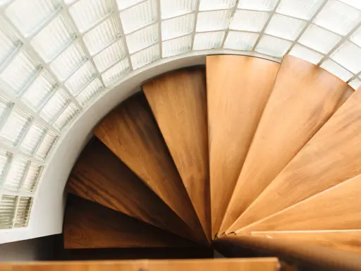 Escalera helicoidal de madera con peldaños que giran bajo un techo de bloques de vidrio translúcido.