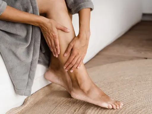 Mani che accarezzano le gambe, evidenziando la pelle secca. La persona è seduta su un letto, avvolta in un accappatoio grigio.