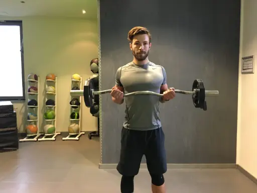 Mann macht Bizeps-Curls mit einer Langhantel. Im Hintergrund stehen Medizinbälle.