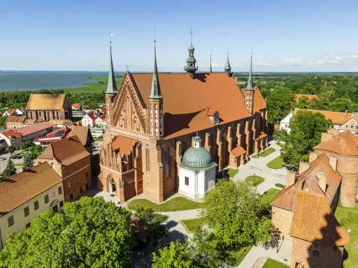 Zamek Frombork z katedrą i wieżami, otoczony zielenią, nad błękitną wodą.