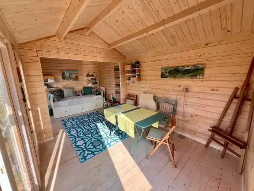 Gemütlicher Gartenhaus Innenausbau mit Tagesbett, Esstisch und Staffelei. Holzwände und Decke schaffen eine warme Atmosphäre.