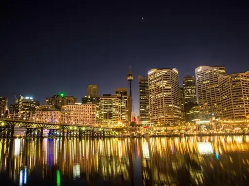 Sydney nocą, z mostem i odbijającymi się w wodzie światłami. Zastanawiasz się, ile się leci do Australii, patrząc na ten widok?