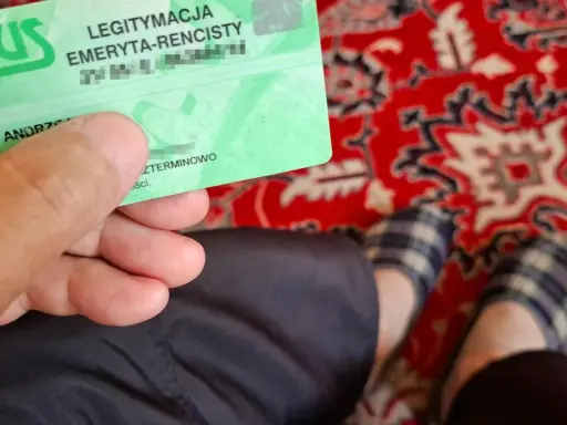Zielona legitymacja ZUS "LEGITYMACJA EMERYTA-RENCISTY" w dłoni. Na dywanie widać stopy w kapciach.