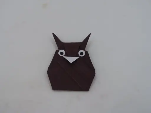 Eine braune origami Eule mit großen weißen Augen und einem kleinen Schnabel sitzt auf hellem Untergrund.