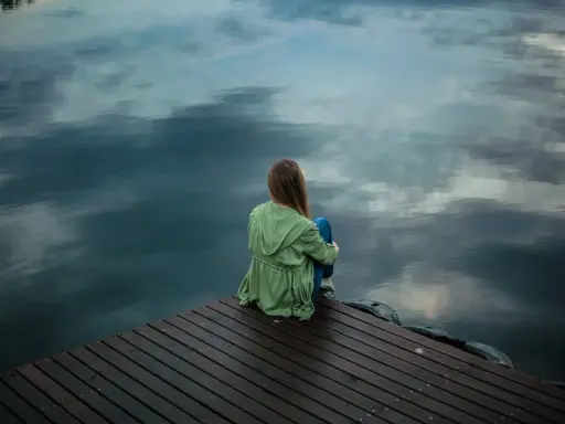 Eine Frau sitzt allein auf einem Steg, blickt aufs Wasser. Die Spiegelung der Wolken im Wasser verstärkt das Gefühl der **Angst vor dem Alleinsein**.