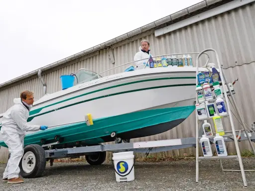 Un homme en combinaison blanche nettoie le flanc d'un bateau blanc et vert sur une remorque. Le nettoyage du pont du bateau est en cours.