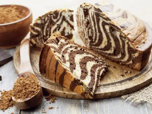 Wilgotne ciasto zebra z czekoladowymi i waniliowymi pasami, pokrojone na plastry, obok kakao i czekolady.