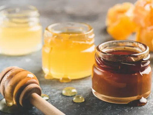 Trois pots de miel aux teintes variées, du clair au foncé, et une cuillère en bois dégoulinante. Quel miel pour pain d'épices ? Le choix est là !