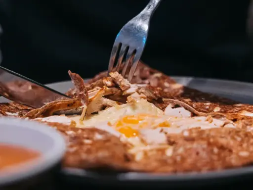 Une galette garnie d'œuf et de poulet, un délice des spécialités de Bretagne, est coupée avec un couteau et une fourchette.