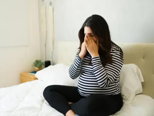Ciężarna kobieta siedzi na łóżku, masując skronie. Dziwny, metaliczny posmak w ustach w ciąży nie daje jej spokoju.