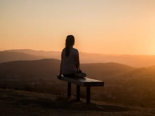 Eine Person sitzt auf einer Bank und blickt in die Ferne. Die untergehende Sonne taucht die Hügellandschaft in warmes Licht, ein Moment, um im Reinen zu sein.