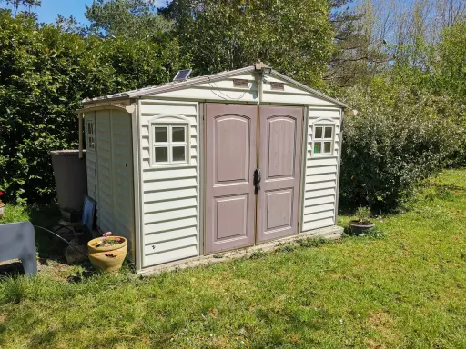 Un abri de jardin en plastique beige, avec des fenêtres et une double porte, est prêt pour son aménagement. Un petit panneau solaire est installé sur le toit.