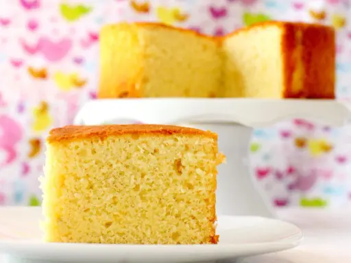 Un delicioso bizcocho de yogur, esponjoso y dorado, servido en un plato blanco. Otro trozo se ve en un soporte para pasteles al fondo.