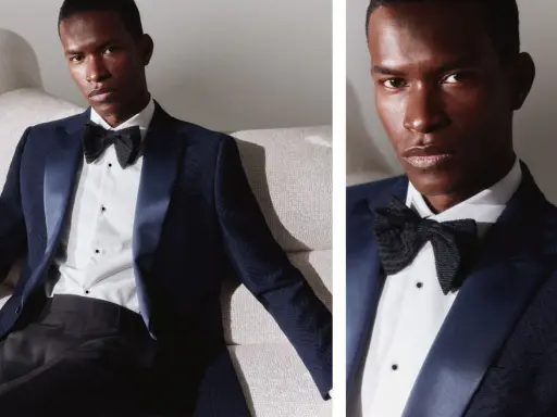 A man in a dark blue dinner jacket vs tuxedo, with a satin lapel and black bow tie, poses on a textured sofa.