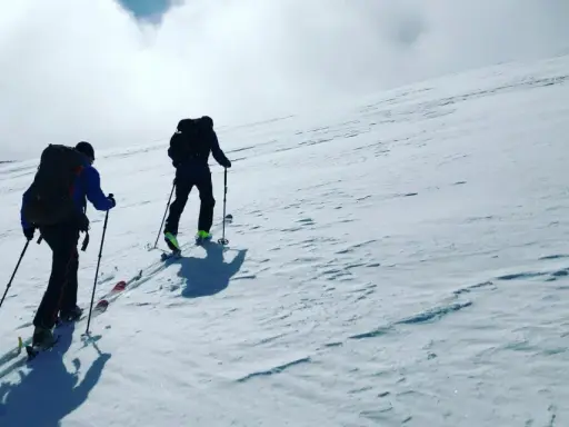 Två personer vandra på vintern uppför en snöklädd sluttning med skidutrustning och ryggsäckar.