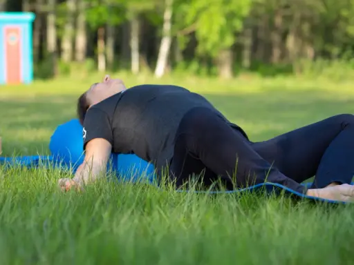 Person ligger på en blå yogarulle i gräset, med böjda knän och armarna utåt. En lugn stund med yin yoga utomhus.