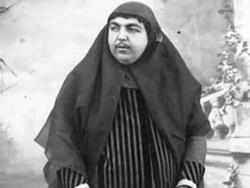 Zahra Khanom Tadj es-Saltaneh, księżniczka perska, w czarnym hidżabie i pasiastym stroju, pozuje na tle ogrodu.