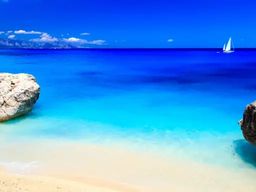 Spiaggia con rocce e mare cristallino, un veliero solca le acque turchesi. Potrebbe essere il mare più bello in Italia.