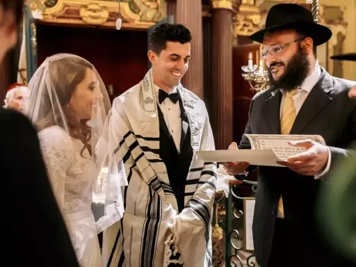 Novios sonríen durante su **matrimonio judío**. El novio lleva un talit y el rabino lee un documento.