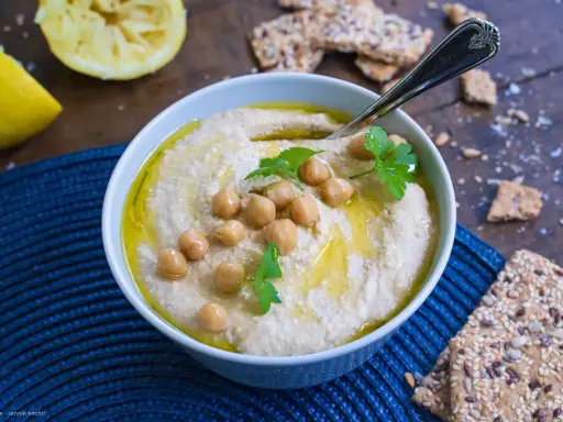 Cremiger Hummus, garniert mit Kichererbsen und Petersilie, daneben Zitronenspalten und knusprige Cracker. Das ist das beste Hummus Rezept!