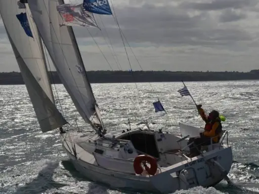 Voilier naviguant par gros temps, le skipper tient un drapeau. La fiche technique du premier 260 est visible sur la coque.