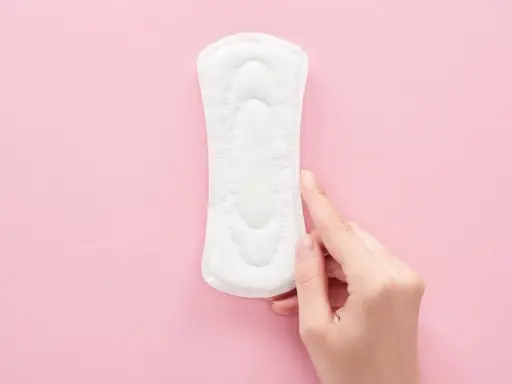 Una mano toca una compresa sobre fondo rosa. Representa cómo es el flujo antes de la regla, ligero y discreto.