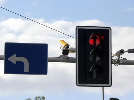 Rejestrator przejazdu na czerwonym świetle, widoczny na tle nieba, wraz z sygnalizacją świetlną i znakiem nakazu skrętu w lewo.