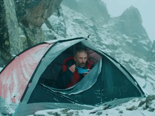 Mężczyzna w namiocie, jedzący posiłek, w zimowej scenerii górskiej. Scena jak z filmów wspinaczkowych.