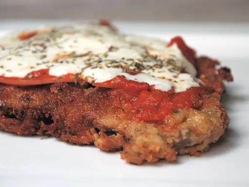 En närbild på en gyllene milanesa napolitana med smält ost, tomatsås och örter.