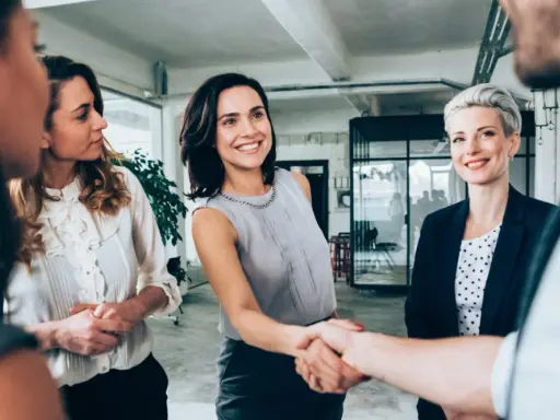 Uśmiechnięte kobiety w biurze, jedna z nich ściska dłoń partnerowi, symbolizując udaną współpracę w spółce partnerskiej.