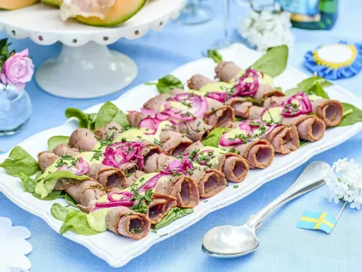 Rullad rostbiff med grön sås och rödlök, perfekt som enkel buffémat till många. Serveras på en vit bricka med gröna blad.
