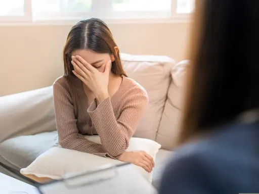 Młoda kobieta siedzi na kanapie, zakrywając twarz dłońmi. Wygląda na apatyczną, jakby straciła wszelką nadzieję.