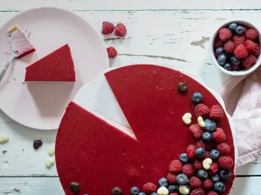 Ein köstlicher no bake Himbeer Cheesecake, dekoriert mit frischen Beeren und Schokoharz. Ein Stück ist bereits auf einem Teller.
