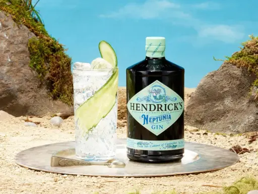 Ein erfrischender Gin Tonic mit Hendricks Neptunia Gin, Gurkenscheiben und Eis, auf sandigem Untergrund mit Felsen und blauem Himmel.