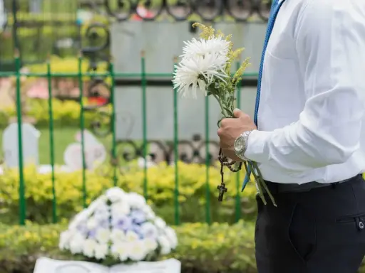 Mężczyzna w białej koszuli i niebieskim krawacie trzyma bukiet białych kwiatów i różaniec. To jego urlop okolicznościowy śmierć bliskiej osoby.