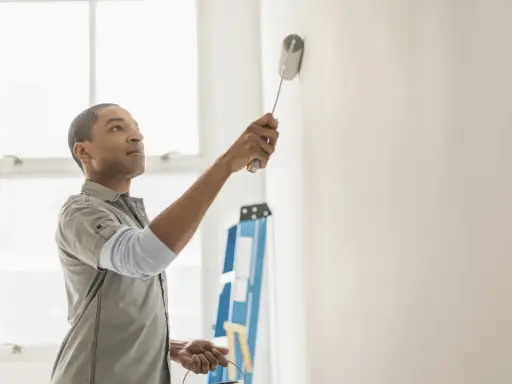 Hombre pintando pared blanca, resolviendo quién paga la pintura en un piso de alquiler. Escalera azul al fondo.