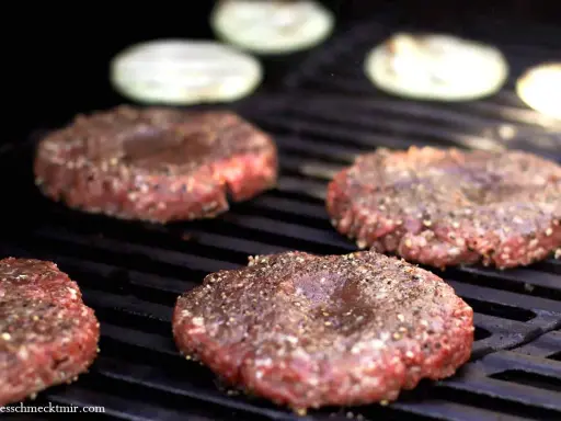 Vier frisch geformte Patties, gewürzt mit Pfeffer, brutzeln auf dem Grill. Perfekt, um Patties selber machen.