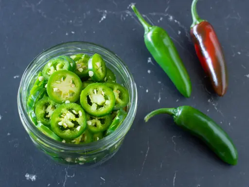 Frisch geschnittene Jalapeños in einem Glas, bereit zum Jalapenos einlegen. Daneben liegen drei ganze Chilis, eine davon reif rot.
