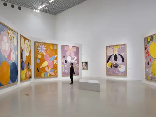En person betraktar färgglada, abstrakta målningar av Hilma af Klint i ett ljust galleri.