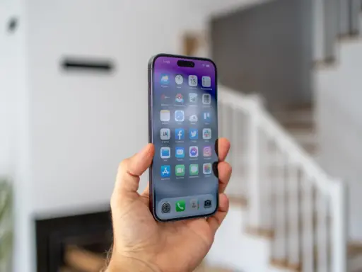 Dłoń trzyma smartfon z widocznym ekranem iPhone 14 Pro Max. Na wyświetlaczu widać ikony aplikacji i godzinę 11:02.