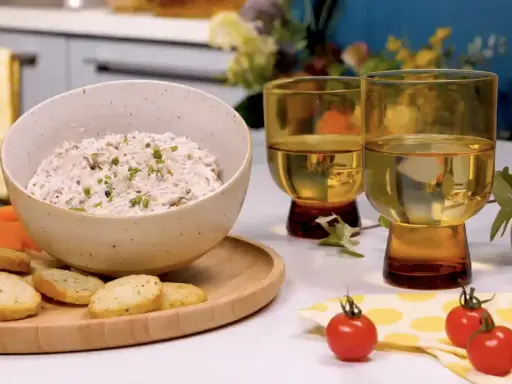 Un bol de rillettes de thon crémeuses, garni de ciboulette, servi avec des crackers, des bâtonnets de carottes et de céleri. Deux verres de vin blanc complètent cette scène gourmande.