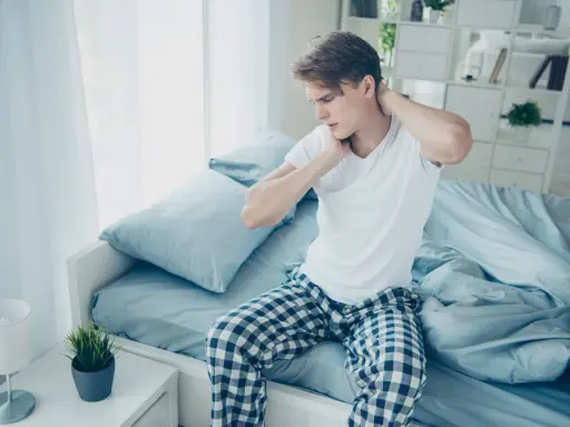 Junger Mann sitzt auf dem Bett und reibt sich den Nacken. Er hat nackenschmerzen nach dem schlafen.
