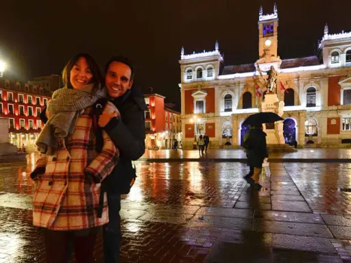 Pareja sonriente en la Plaza Mayor de Valladolid, disfrutando de las atracciones nocturnas y el reflejo de las luces en el suelo mojado.